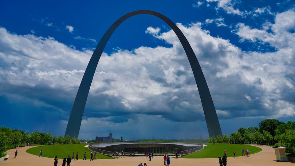 A large arch structure with a circular building at its base, surrounded by green lawns and people.