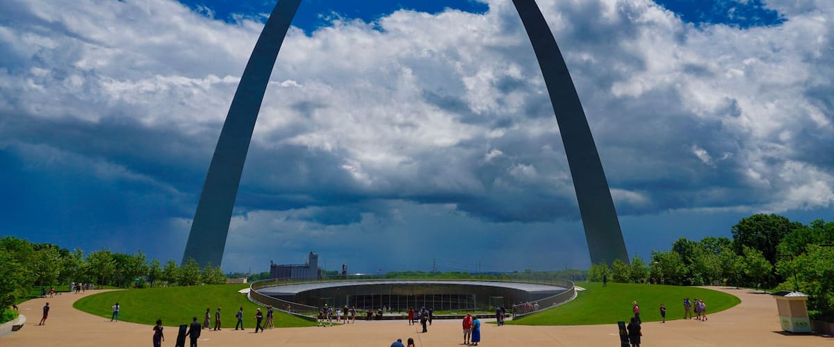 A large arch structure with a circular building at its base, surrounded by green lawns and people.