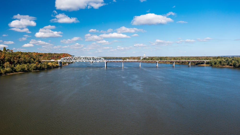 Eine weiße Brücke über einem ruhigen Fluss, im Hintergrund Bäume und Hügel.