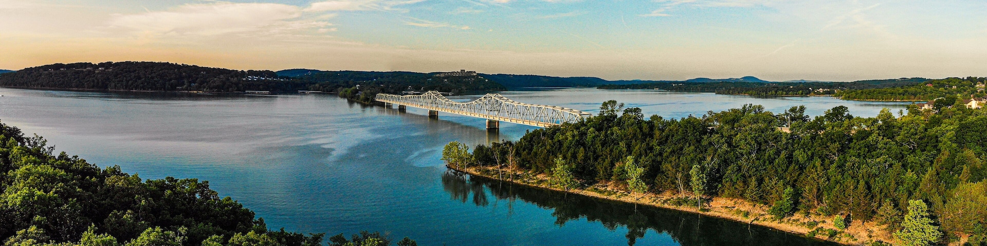 A bridge over a calm river surrounded by dense forests and distant hills.