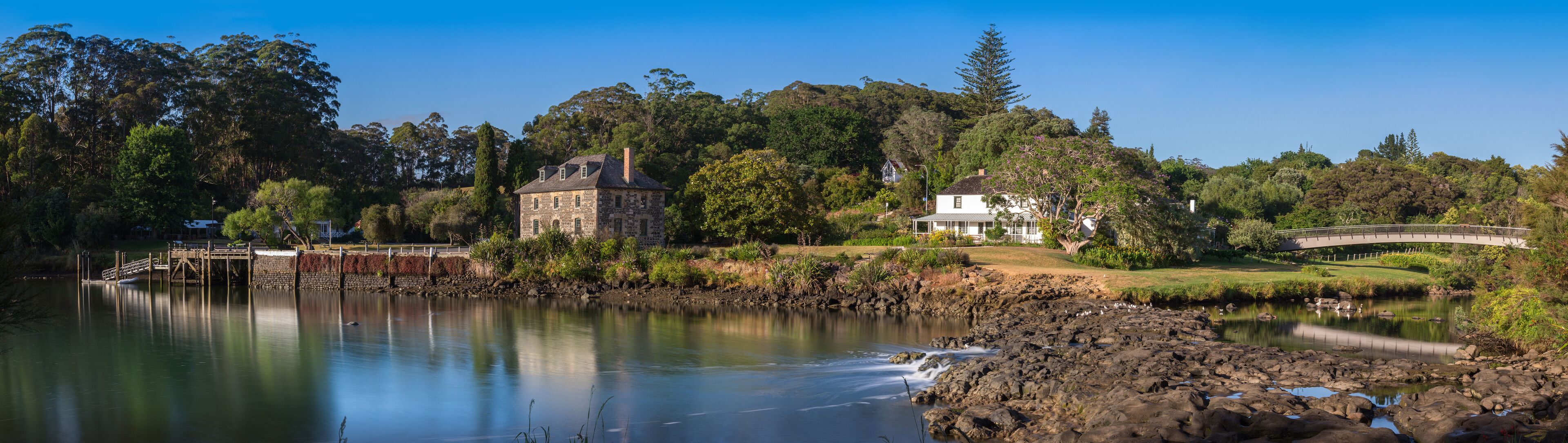 A lake with a stone bridge, a stone house, and a white house surrounded by trees.