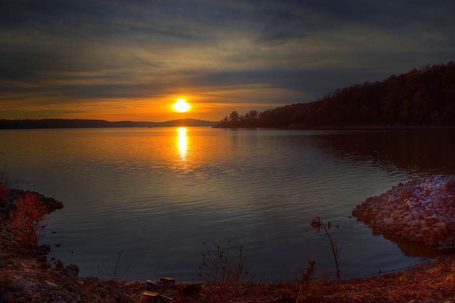 Un atardecer sobre un lago tranquilo con una costa rocosa y árboles al fondo.