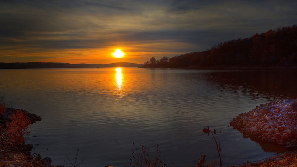 A sunset over a calm lake with a rocky shoreline and trees in the background.