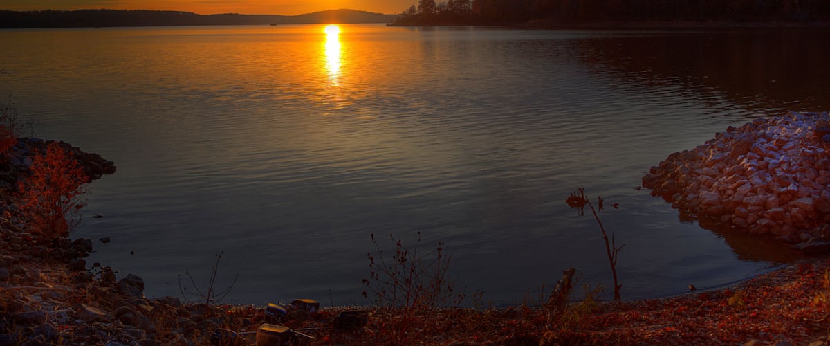 A sunset over a calm lake with a rocky shoreline and trees in the background.