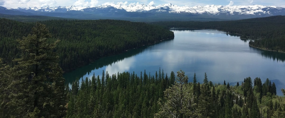 A lake surrounded by dense forests and snow-capped mountains under a partly cloudy sky.