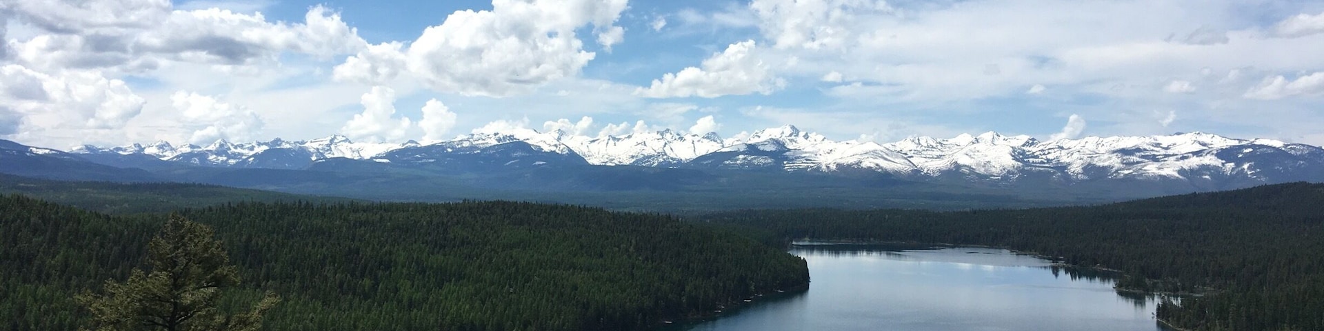 A lake surrounded by dense forests and snow-capped mountains under a partly cloudy sky.