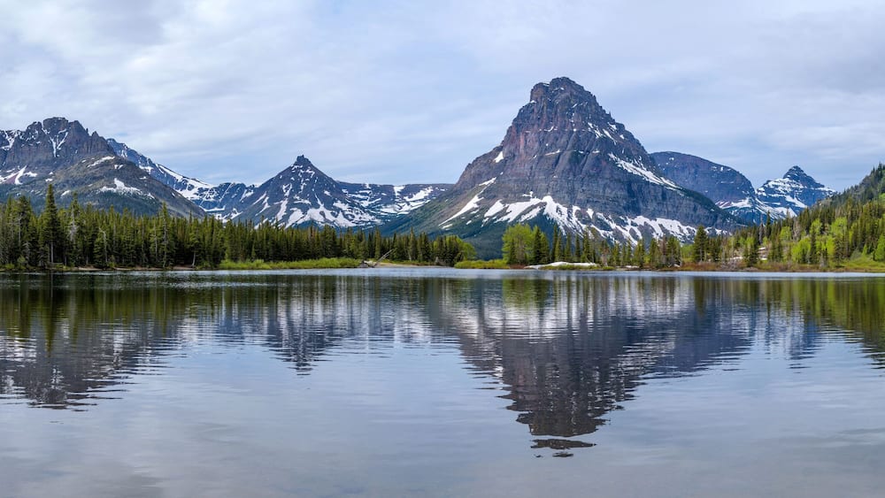 A calm lake reflecting mountains with snow-capped peaks and dense forests.