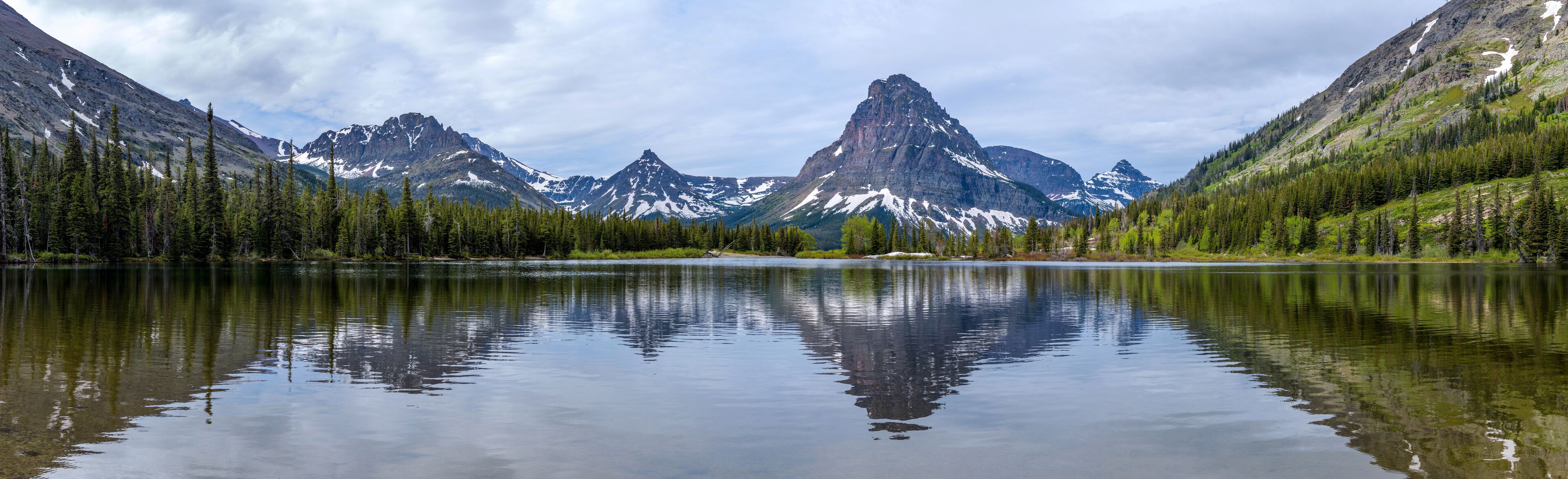 A calm lake reflecting mountains with snow-capped peaks and dense forests.