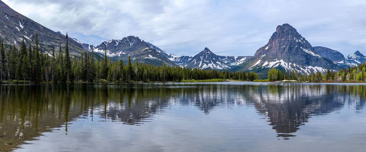 A calm lake reflecting mountains with snow-capped peaks and dense forests.