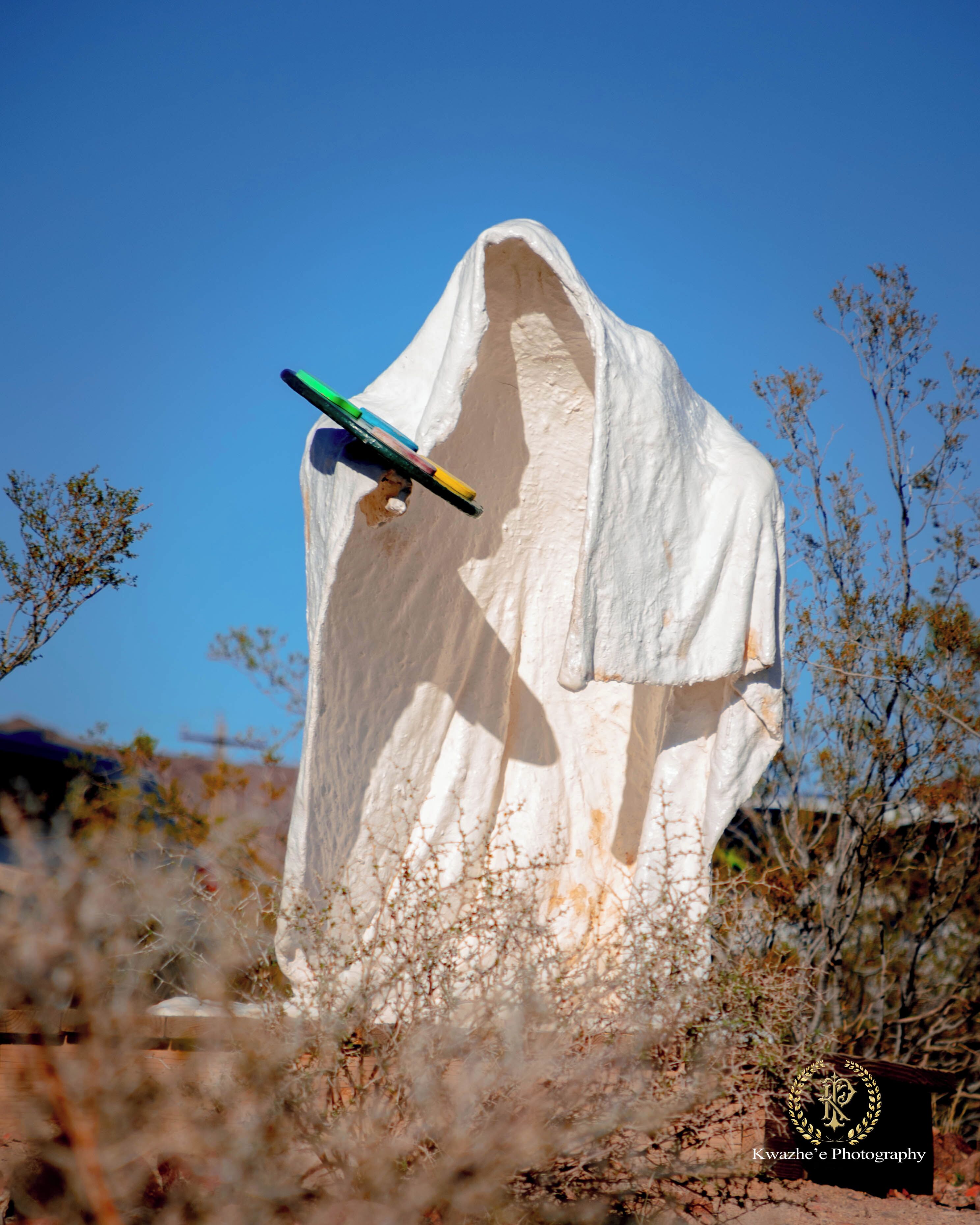 A white ghost-like statue with a colorful object on its head, surrounded by dry vegetation and a clear blue sky.