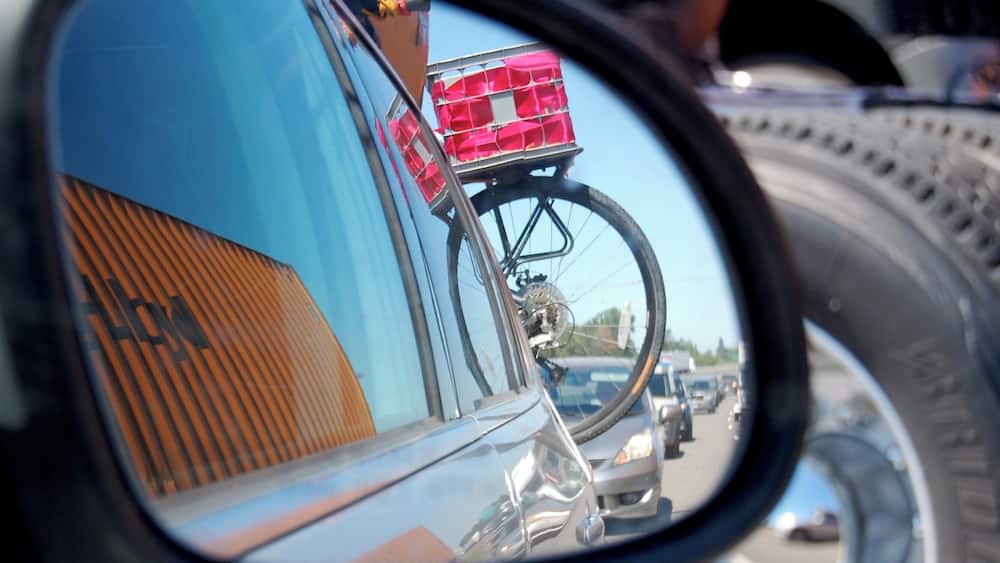 Le reflet dans le rétroviseur d’une voiture montre un vélo avec un panier rose, le rétroviseur d’une autre voiture et un pneu.