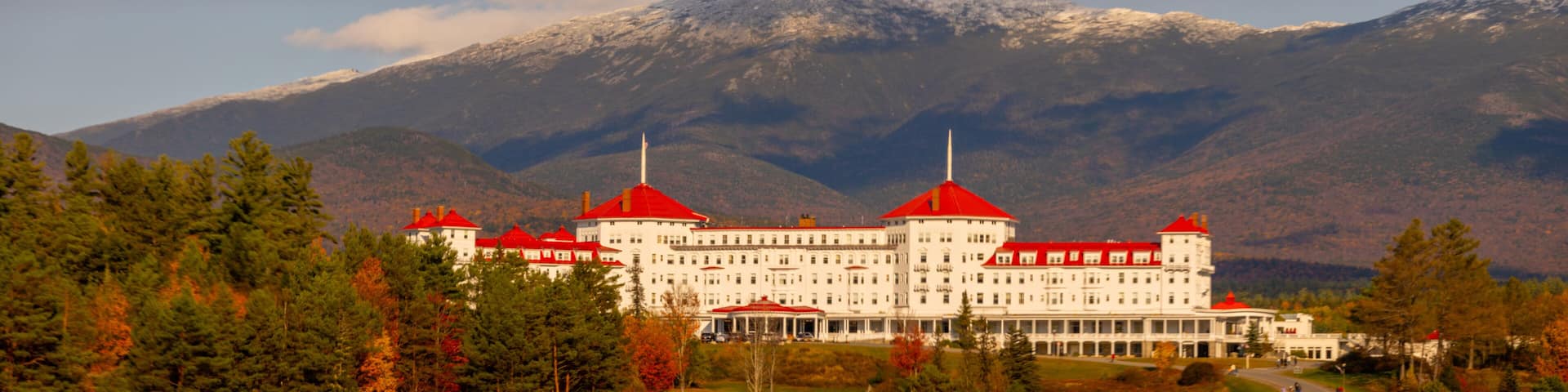 A large white building with red roofs, surrounded by trees and mountains.