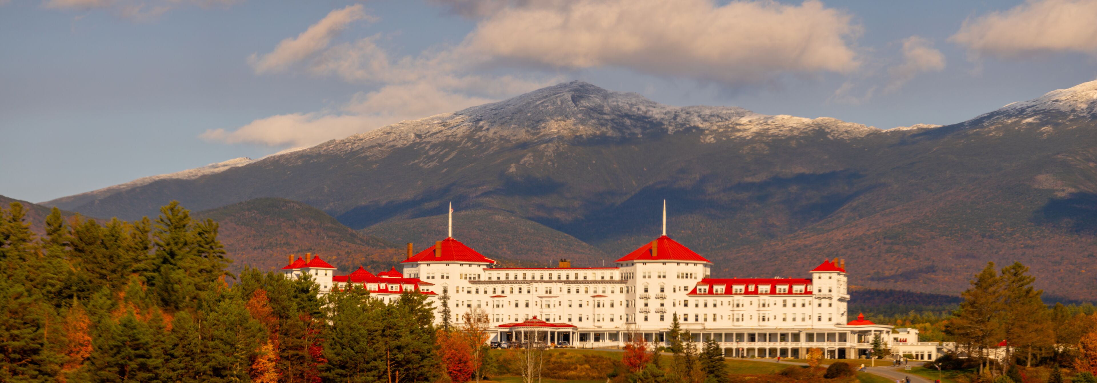 A large white building with red roofs, surrounded by trees and mountains.