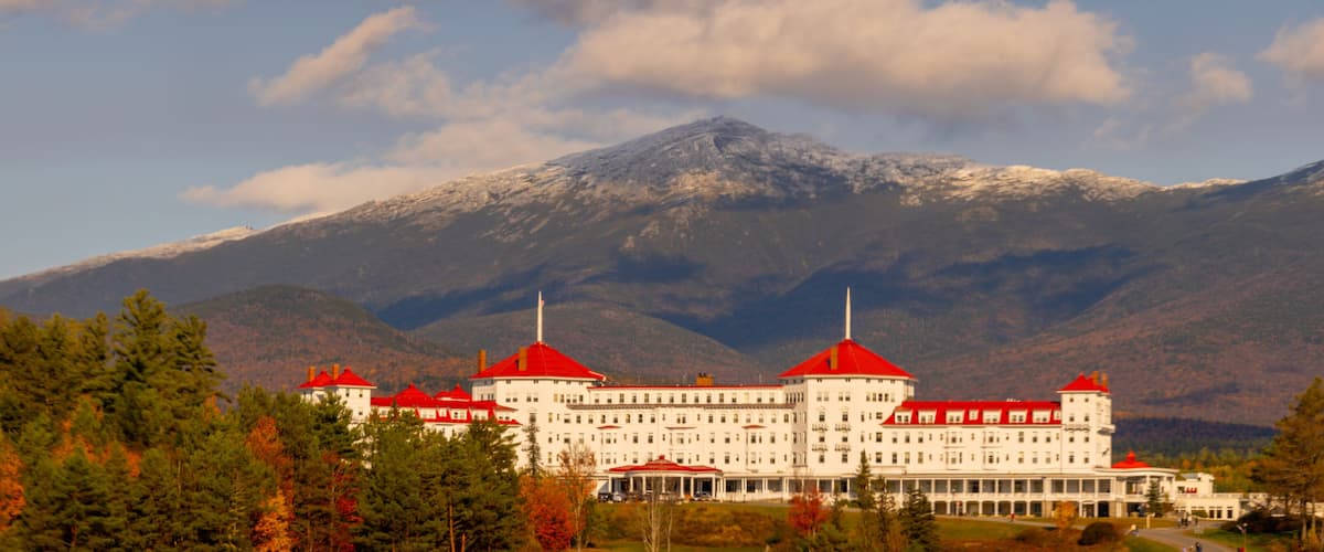 A large white building with red roofs, surrounded by trees and mountains.