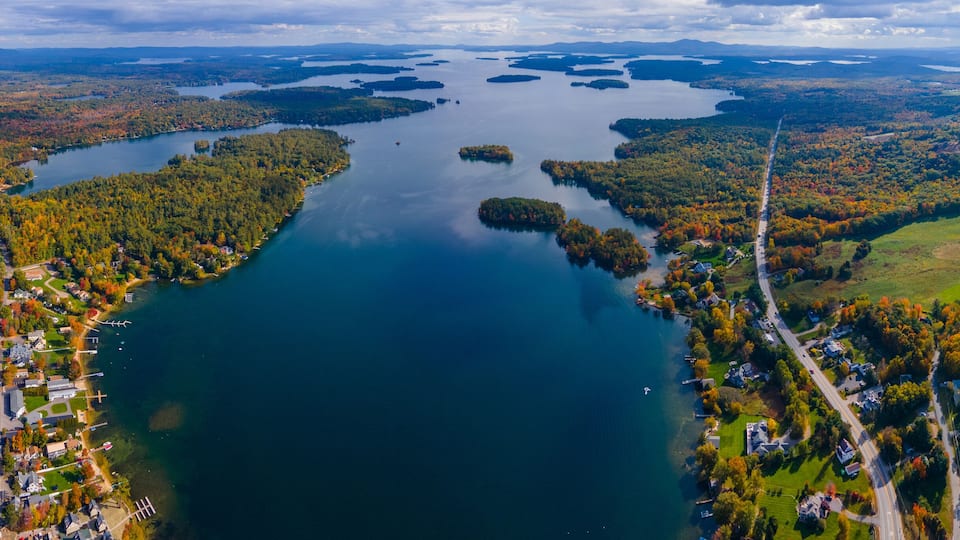 An aerial view of a lake surrounded by autumn-colored forests and residential areas.