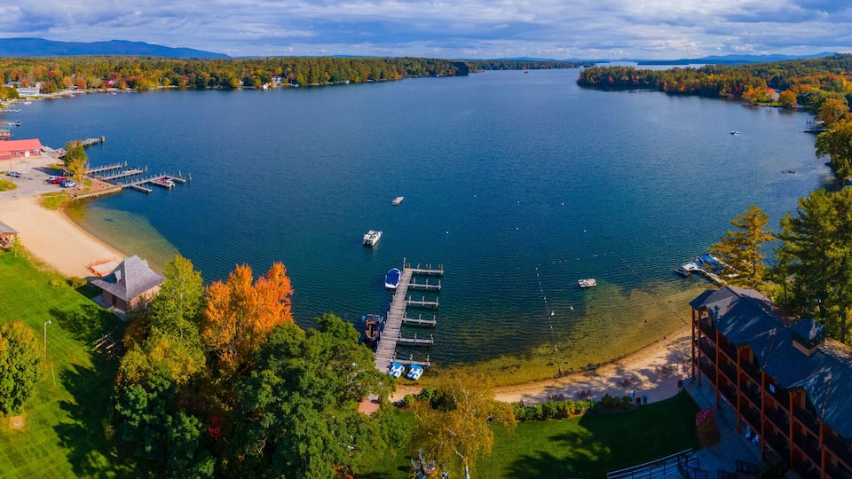A lake surrounded by autumn-colored trees, a dock, and a small town with buildings and a basketball court.