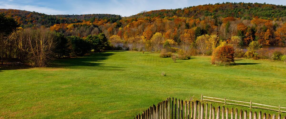 A grassy field with a wooden fence, surrounded by autumn-colored trees and a hill in the background.