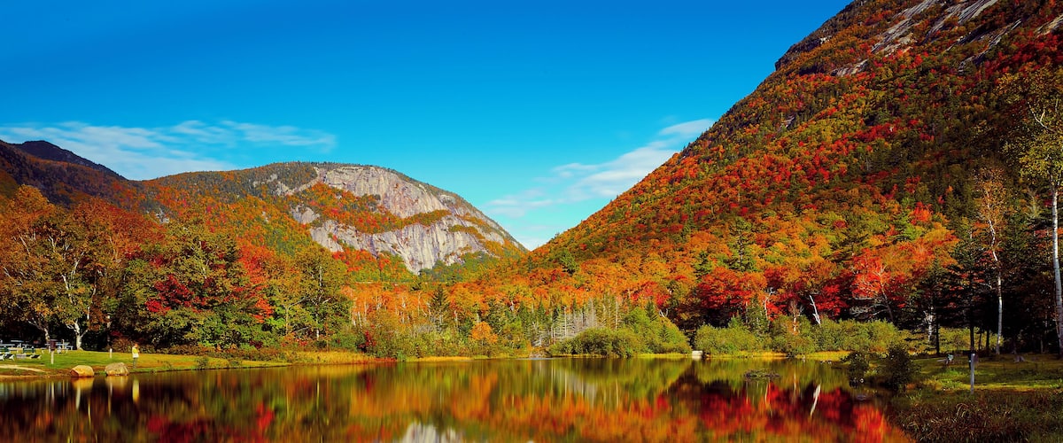 A lake reflecting colorful autumn foliage, surrounded by mountains and a clear blue sky.