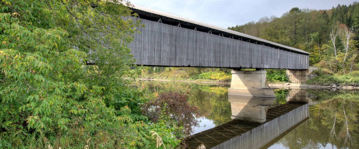 A covered bridge spanning a calm river, surrounded by lush greenery and trees.