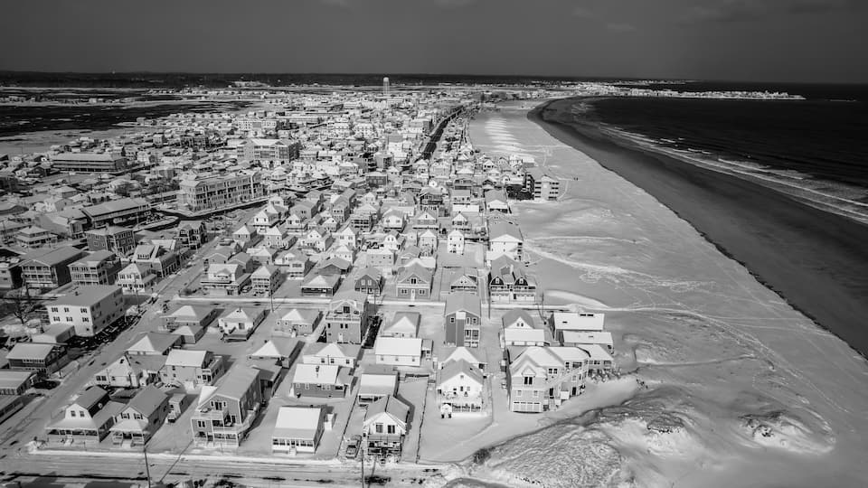An aerial view of a coastal town with a beach, residential buildings, and a lighthouse in the distance.
