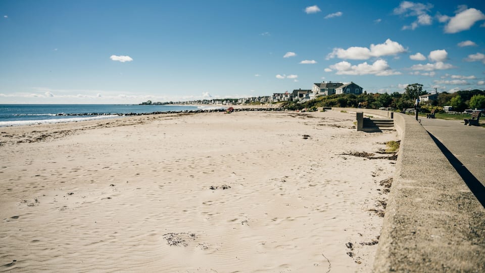 A sandy beach with a concrete walkway, ocean, and houses in the background.