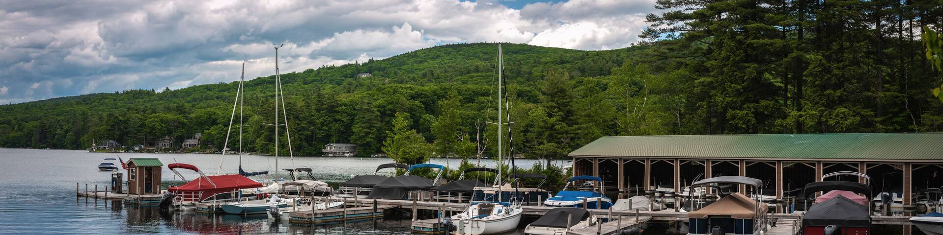 A calm lake with boats docked at a pier, surrounded by lush green hills and a cloudy sky.