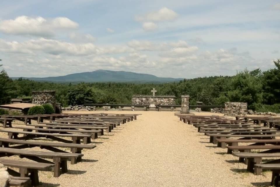 Un chemin de gravier bordé de bancs en bois mène à une structure en pierre surmontée d’une croix, le tout étant entouré de verdure et avec des montagnes visibles au loin.