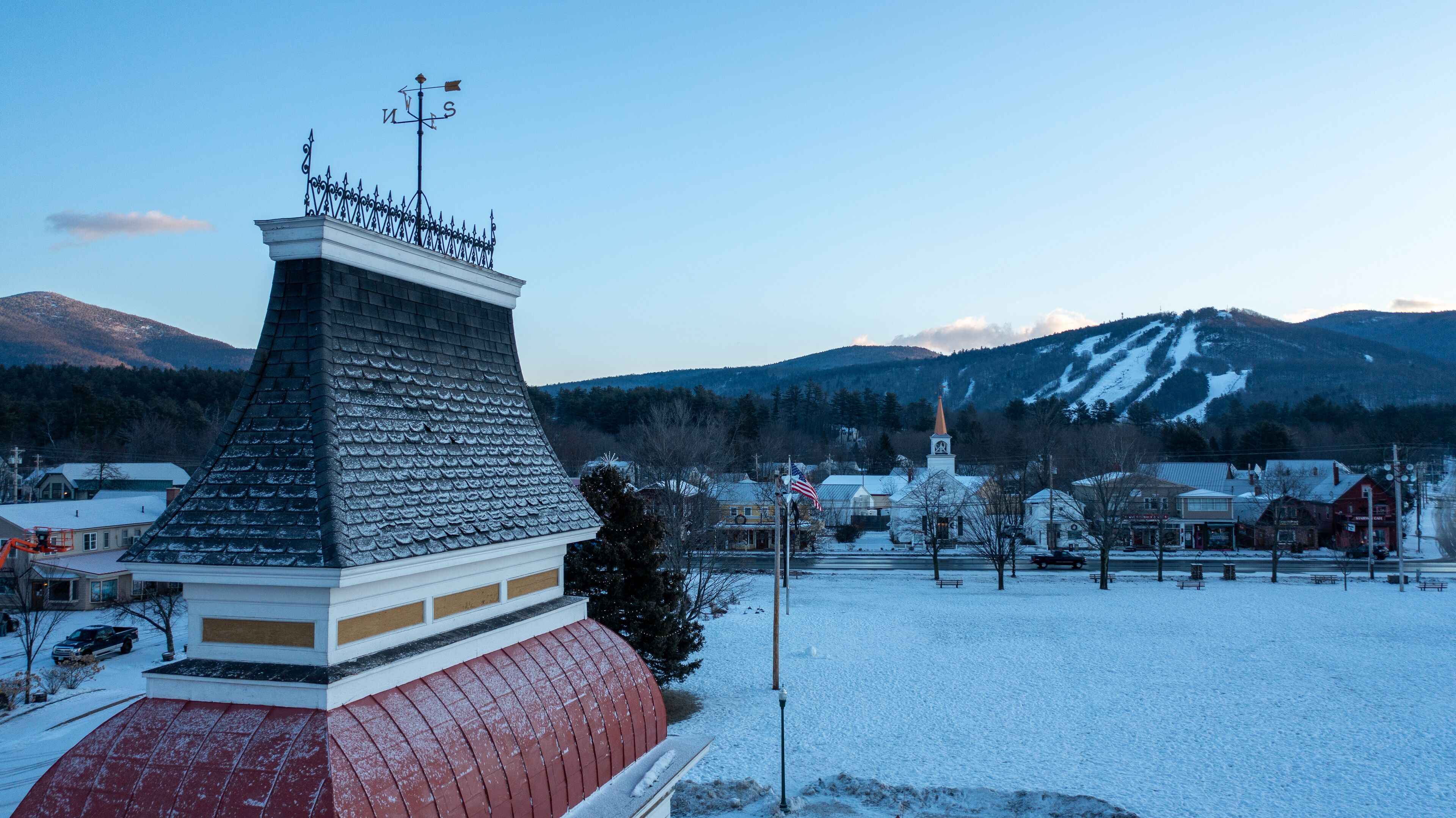 A snow-covered town with a church steeple, a red-roofed building, and mountains in the background.