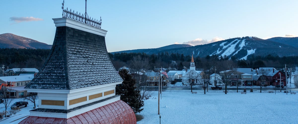 A snow-covered town with a church steeple, a red-roofed building, and mountains in the background.