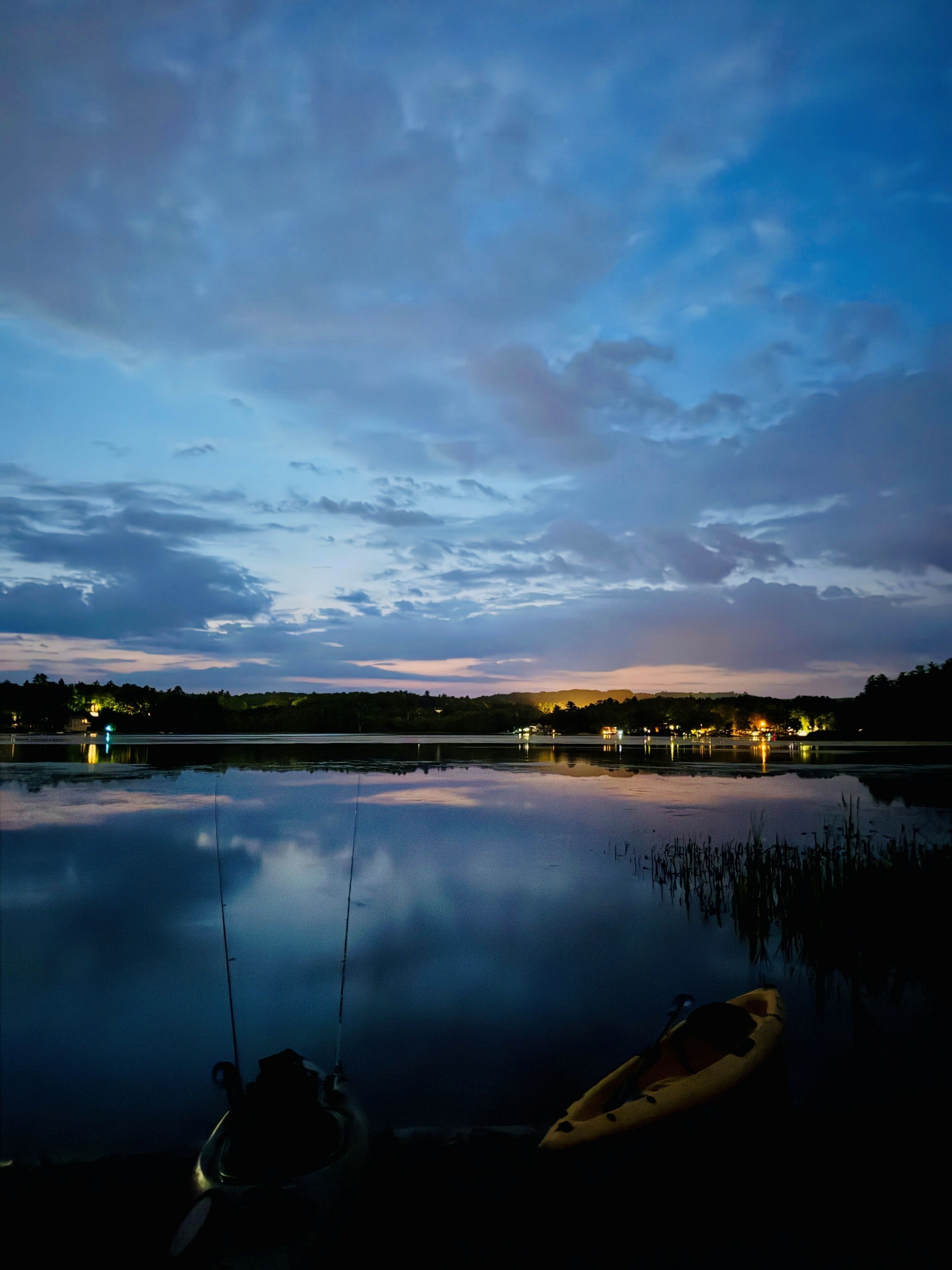 Un lago sereno al anochecer con un kayak y una caña de pescar en primer plano, y una orilla distante con luces.