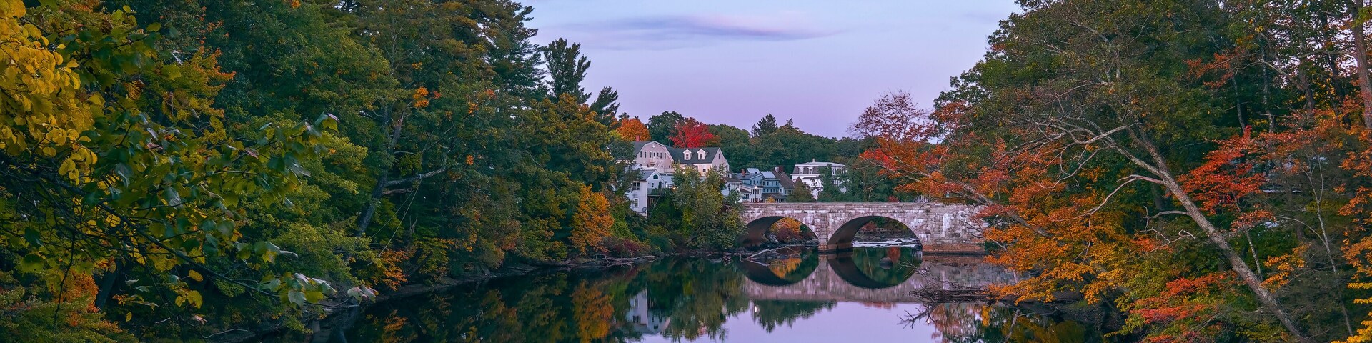 Un lago sereno con follaje otoñal, un puente de piedra y casas al fondo.