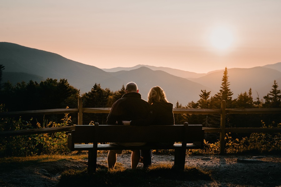 A couple sitting on a bench overlooking a mountainous landscape during sunset.