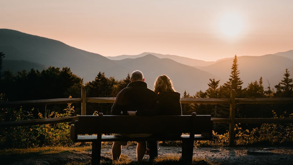 A couple sitting on a bench overlooking a mountainous landscape during sunset.