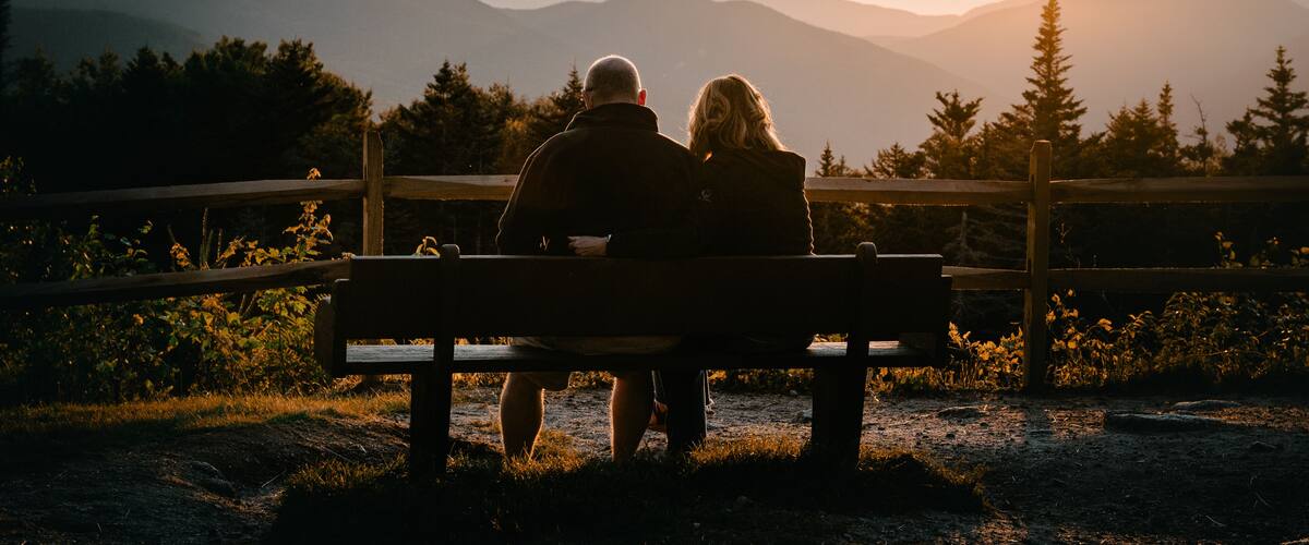 A couple sitting on a bench overlooking a mountainous landscape during sunset.
