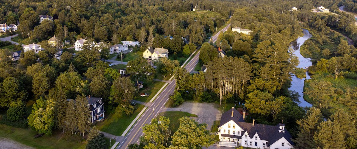 Paisagem rural com um rio, casas e montanhas ao fundo.