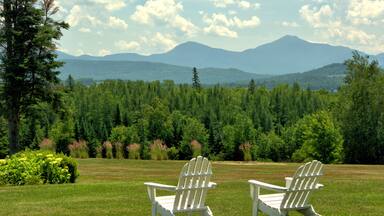 Inviting and restful scenic vista in Whitefield, New Hampshire. White lawn chairs overlooking hillside of lush evergreen trees framed by distant mountains in the White Mountains Presidential Range.