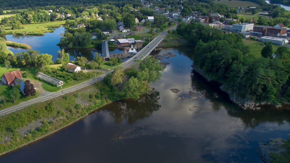 A river flowing through a town with a bridge, surrounded by greenery and mountains.