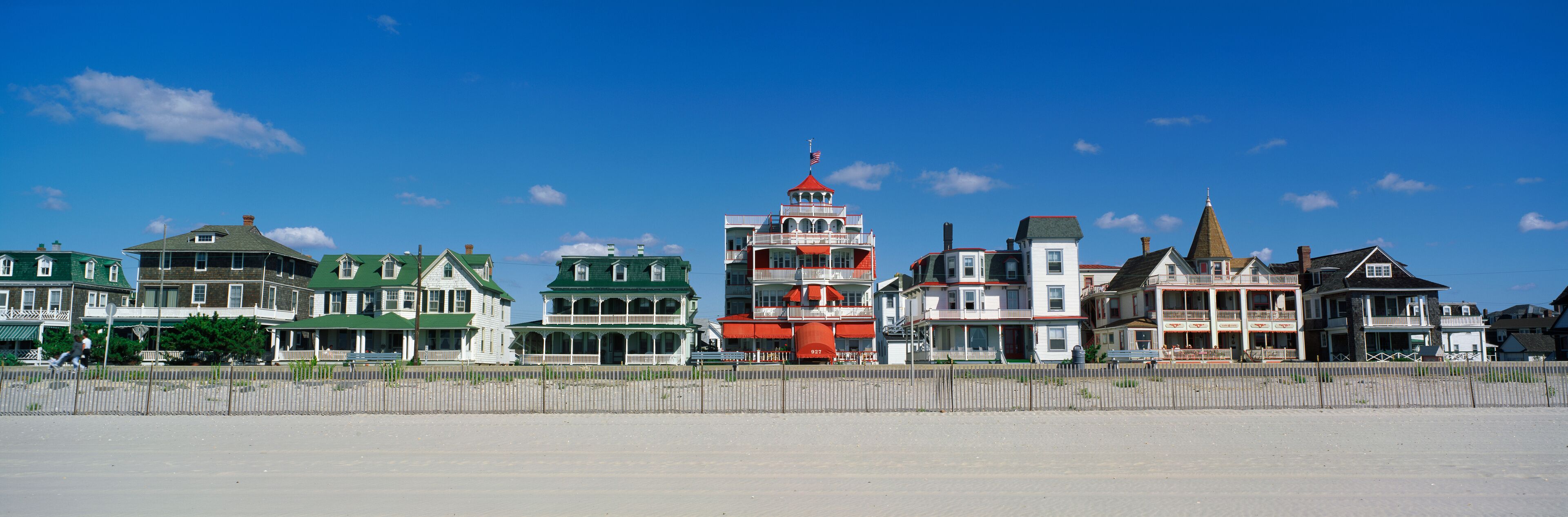 These are Victorian style homes overlooking the beach in Cape May. There is wooden fence separating the beach and the houses. The homes have large front porches. The sky is a deep blue with just a few white puffy clouds.