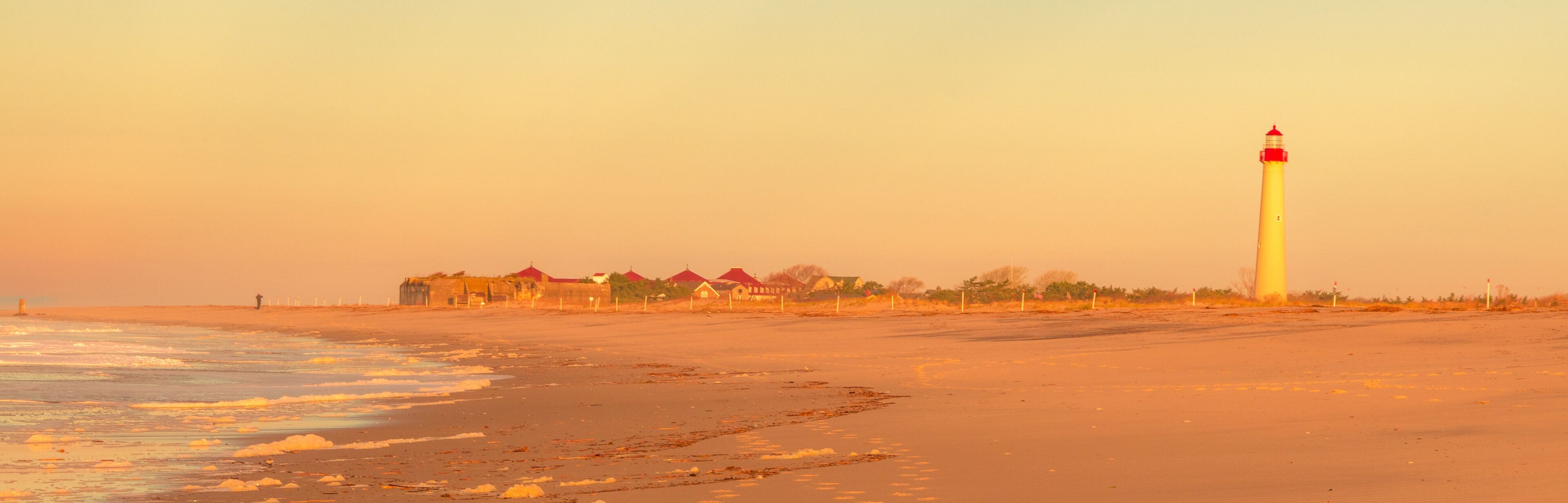 A coastal scene with a lighthouse, beach, and row of houses with red roofs.