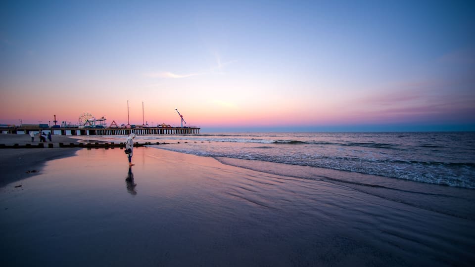 A beach with a pier extending into the water, a Ferris wheel in the background, and a sunset sky.