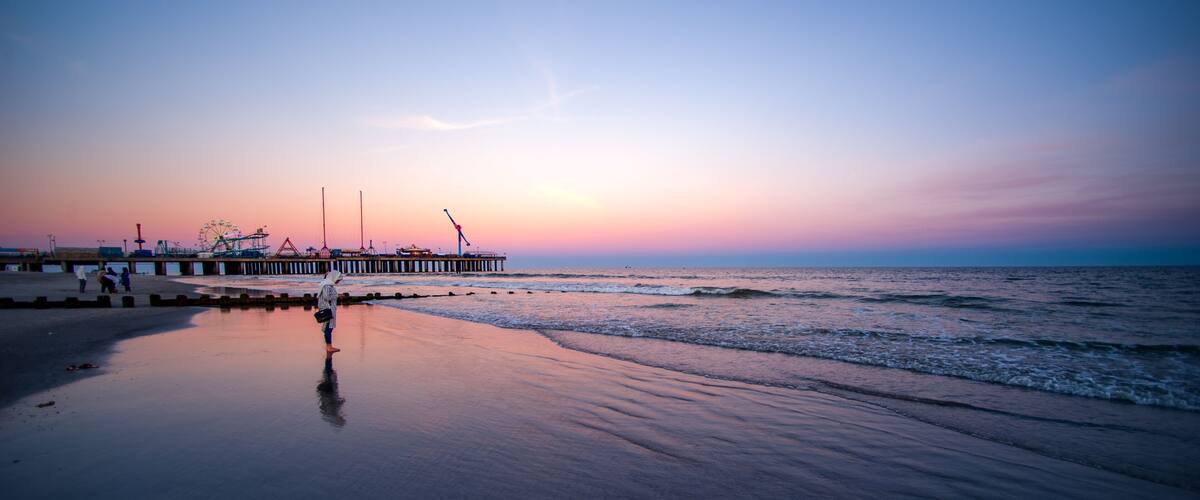 A beach with a pier extending into the water, a Ferris wheel in the background, and a sunset sky.
