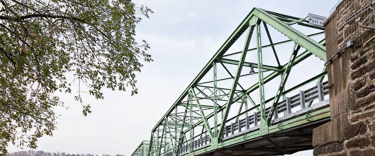 A green metal bridge over a calm river with a stone base, surrounded by trees and a distant hill.