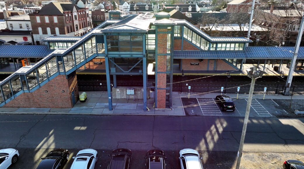 Eine mehrstöckige Fußgängerbrücke mit Glasgeländern und einer Ziegelstruktur, die zwei Gebäude miteinander verbindet, unterhalb befindet sich ein Parkplatz.