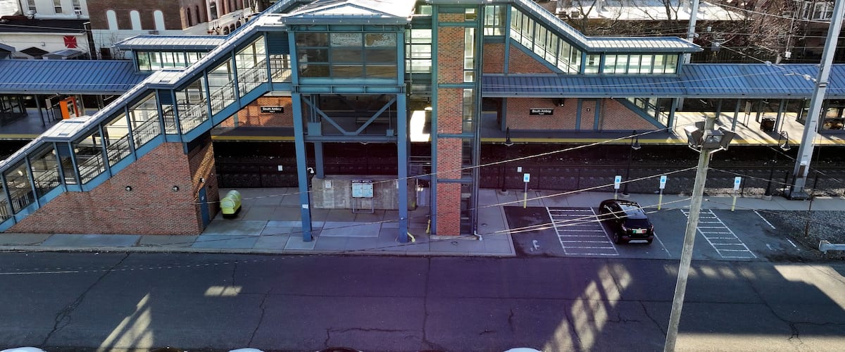 A multi-level pedestrian bridge with glass railings and a brick structure, connecting two buildings with a parking area below.