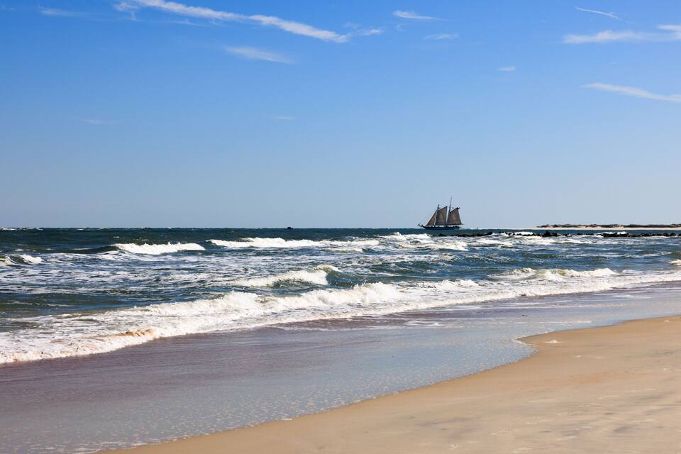 A beach with waves, a sandy shore, and a sailboat in the distance.