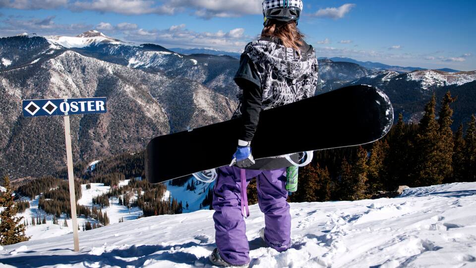 A snowboarder in purple gear standing on a snowy slope with a mountain view in the background.