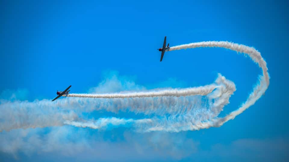 Two airplanes performing aerial maneuvers with smoke trails against a clear blue sky.