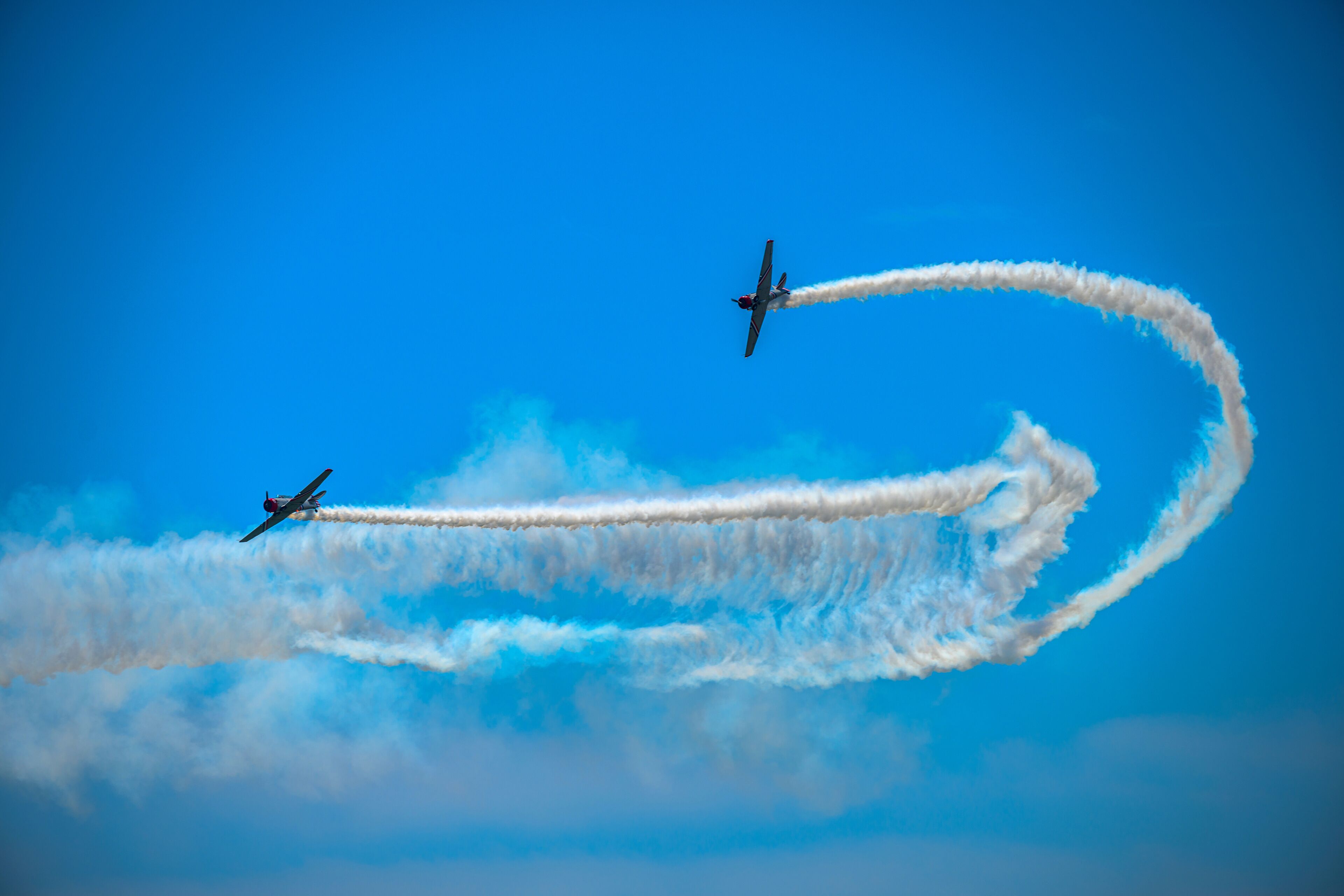 Two airplanes performing aerial maneuvers with smoke trails against a clear blue sky.