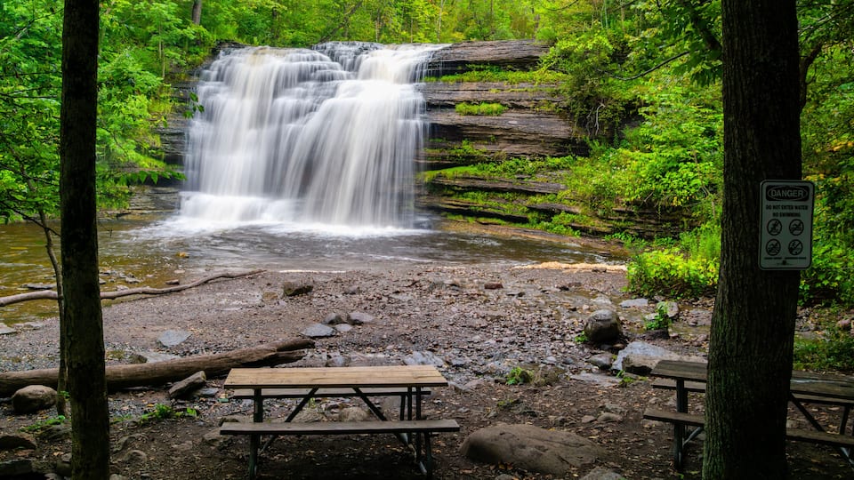 A waterfall in a forested area with picnic tables and a warning sign.
