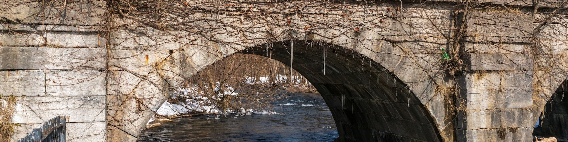 Uma ponte de pedra sobre um rio, com neve no chão e árvores sem folhas.